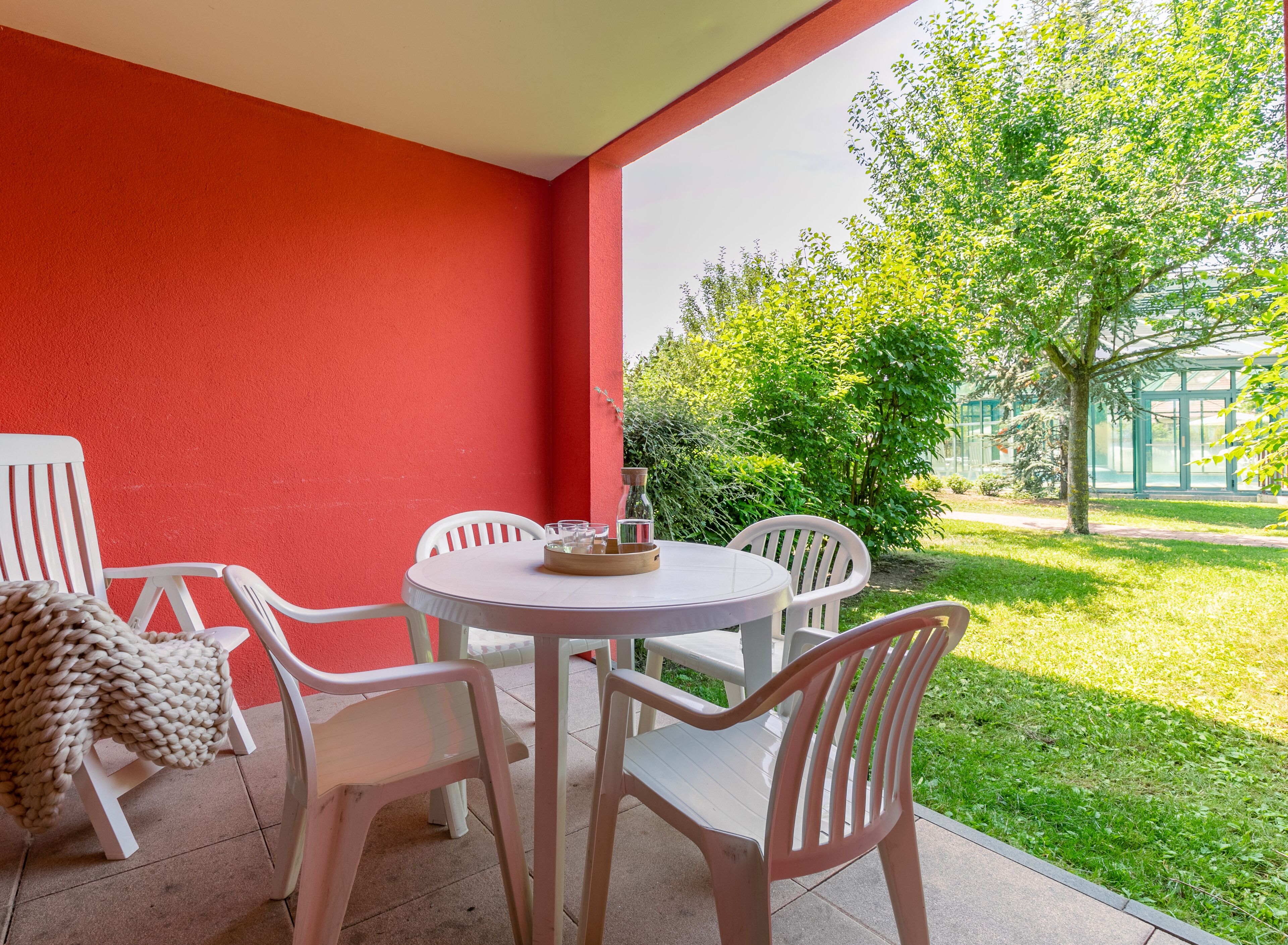 A patio with a round table and white plastic chairs, a red wall, and a view of a green garden.
