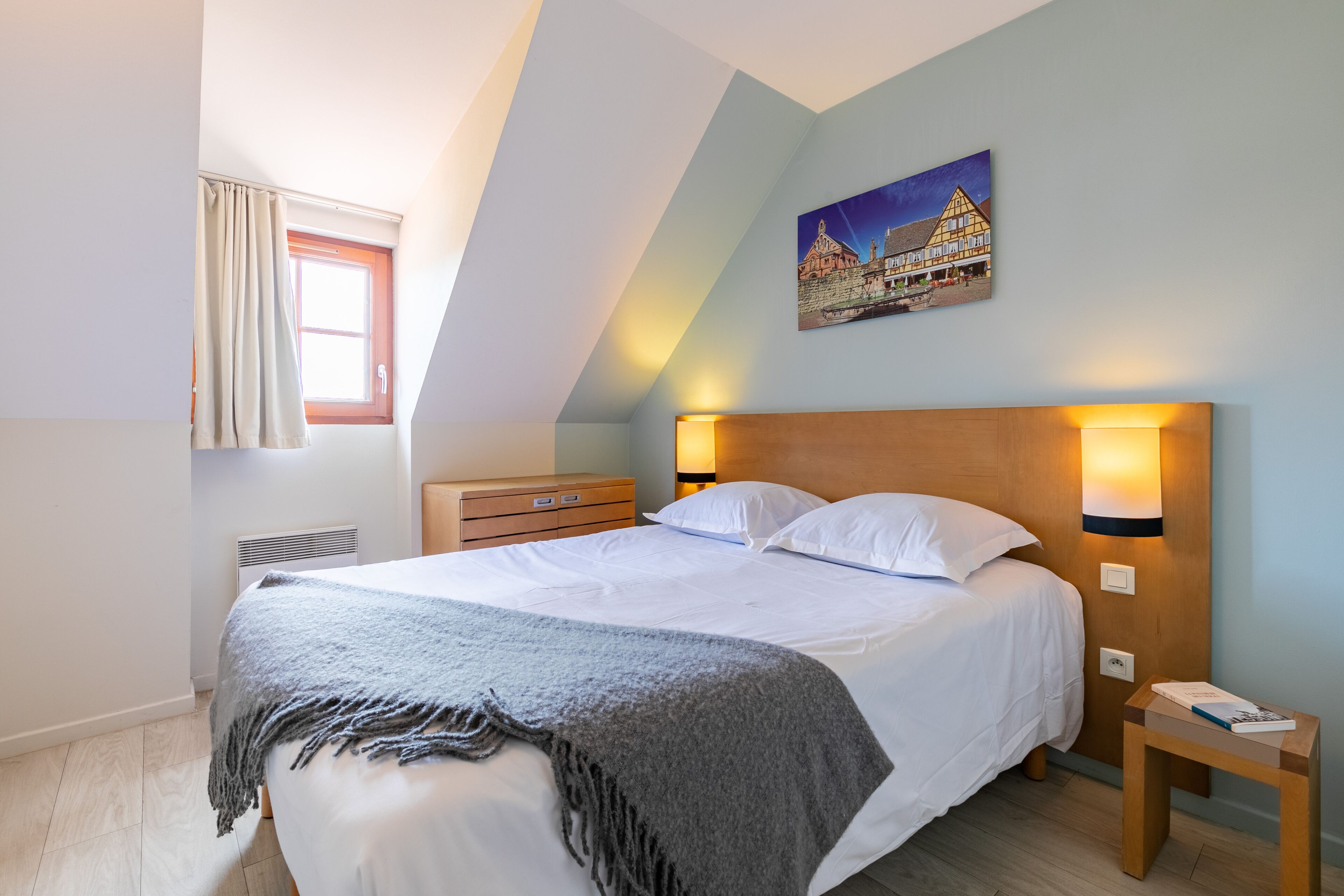 A bedroom with a bed, a nightstand, a lamp, a window with curtains, and a painting on the wall.