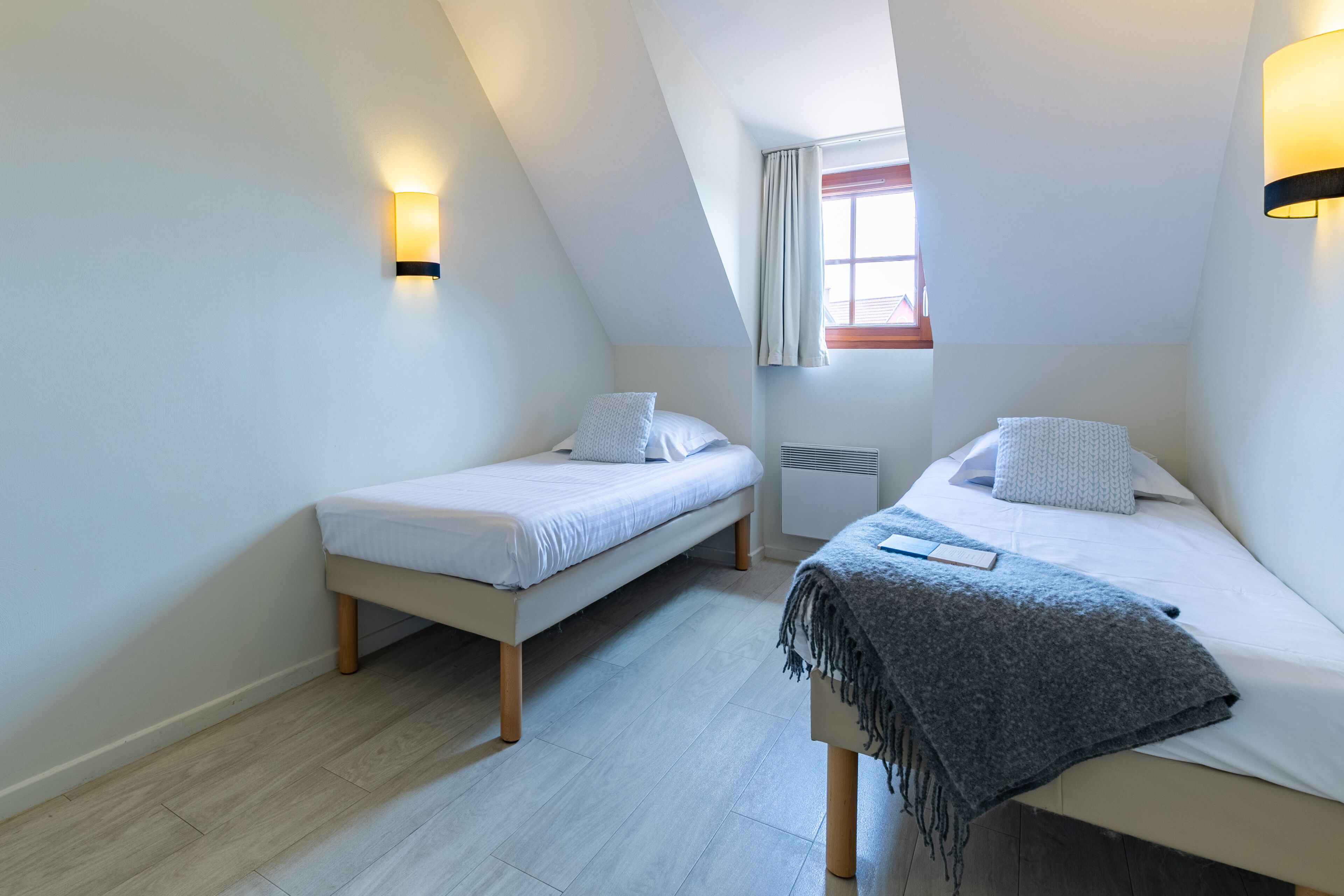 Een kleine, moderne slaapkamer met twee eenpersoonsbedden, een raam met gordijnen en wandlampen.