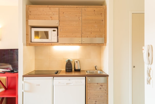 Eine kompakte Küche mit Holzschränken, einer Mikrowelle, einer Kaffeemaschine, einem Geschirrspüler und einem roten Tisch mit einer Fernbedienung.