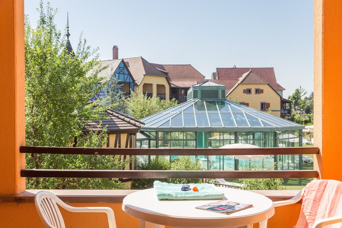 A balcony with a table and chairs, overlooking a garden and buildings with traditional architecture.