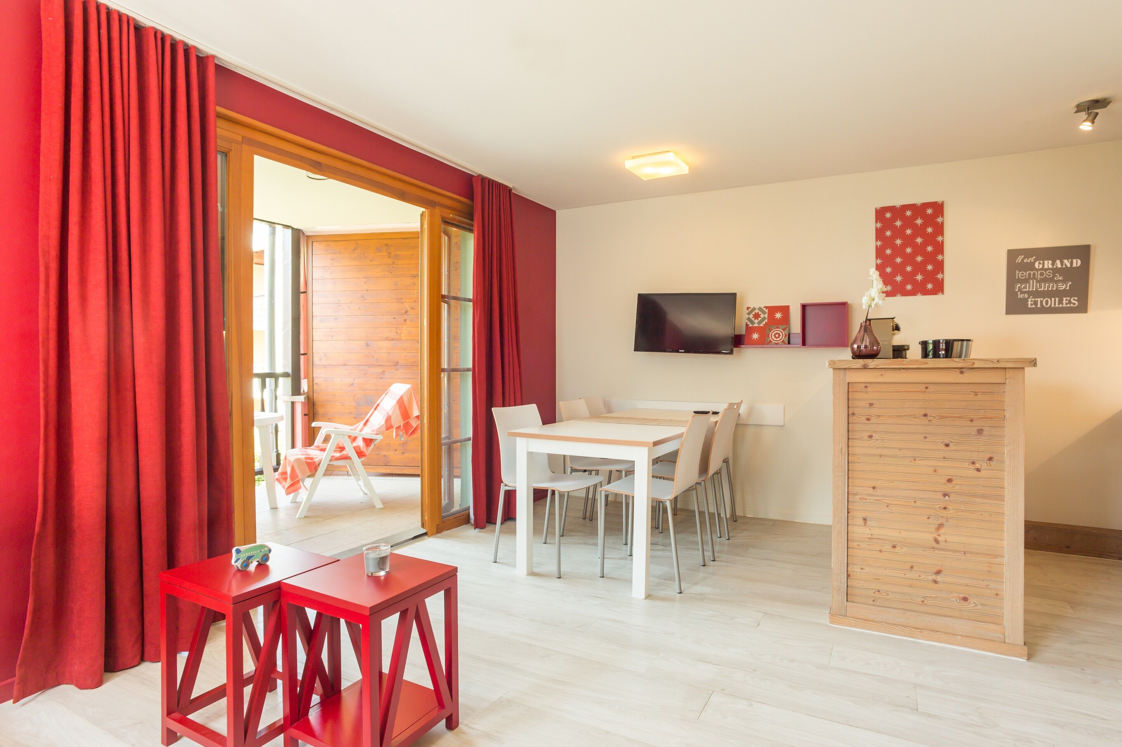 A room with a wooden dining table and chairs, a red table, a TV mounted on the wall, and a wooden cabinet.
