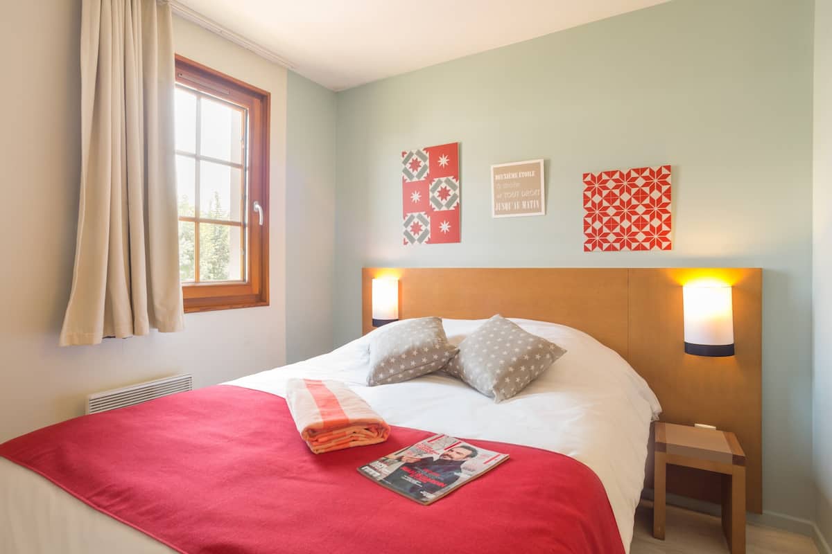 A neatly made bed with a red blanket, pillows, and a folded magazine on it. There are wall-mounted lights, a window with curtains, and a radiator.