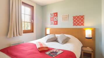 A neatly made bed with a red blanket, pillows, and a folded magazine on it. There are wall-mounted lights, a window with curtains, and a radiator.