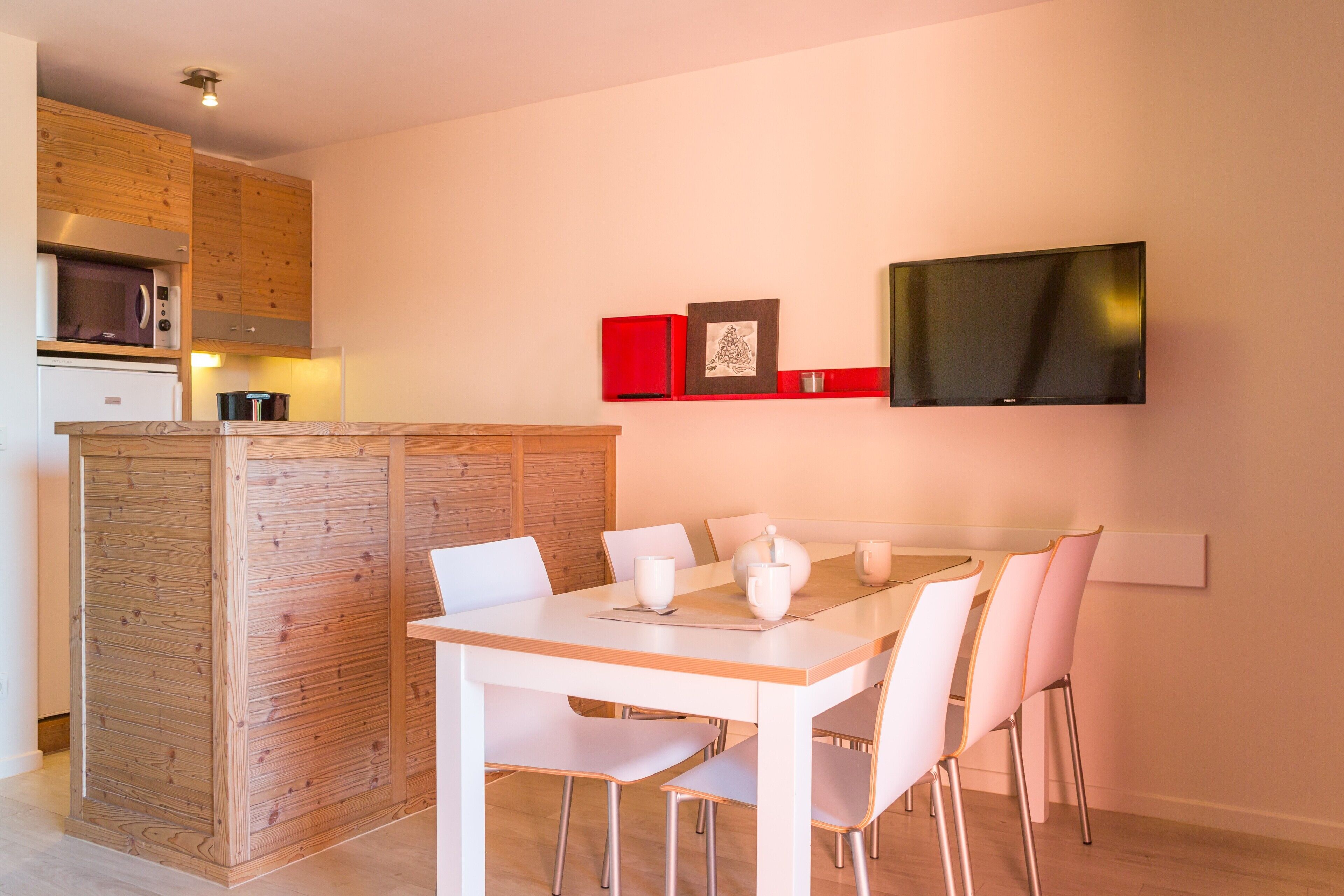 A modern kitchen with wooden cabinets, a microwave, and a stove. There is a dining table with white chairs and a flat-screen TV mounted on the wall.