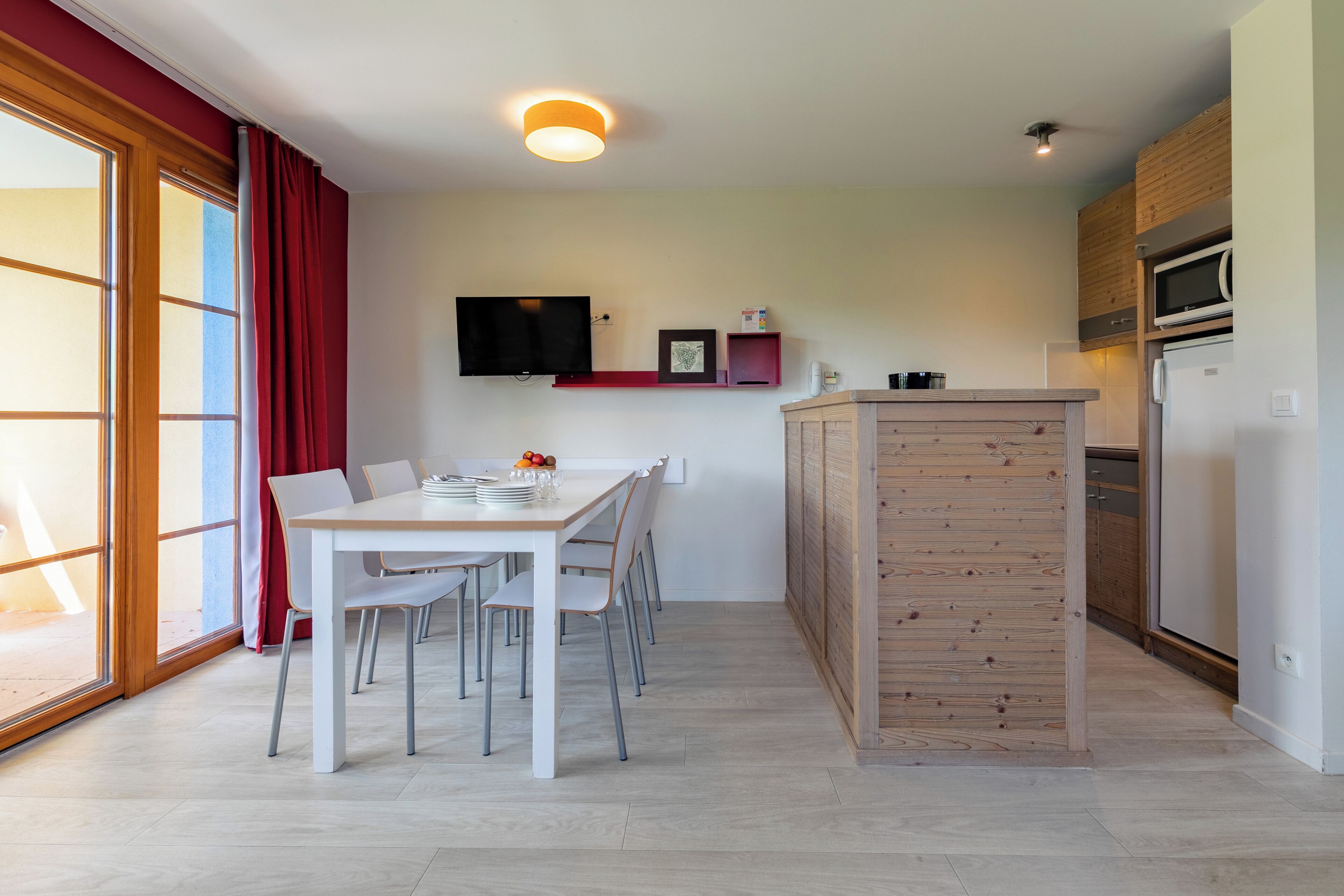 Una cocina moderna con una isla de madera, un comedor con sillas blancas y un amplio ventanal con cortinas rojas.