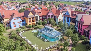 A colorful hotel complex with a central glass-enclosed pool area, surrounded by lush greenery and traditional European architecture. - Pierre & Vacances Résidence Le Clos d'Eguisheim (Eguisheim)