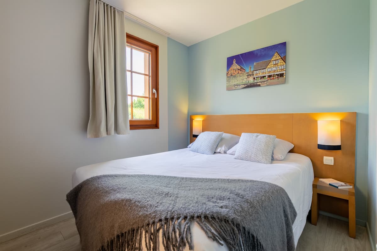 A bedroom with a bed, a nightstand, a lamp, a window with curtains, and a painting on the wall.