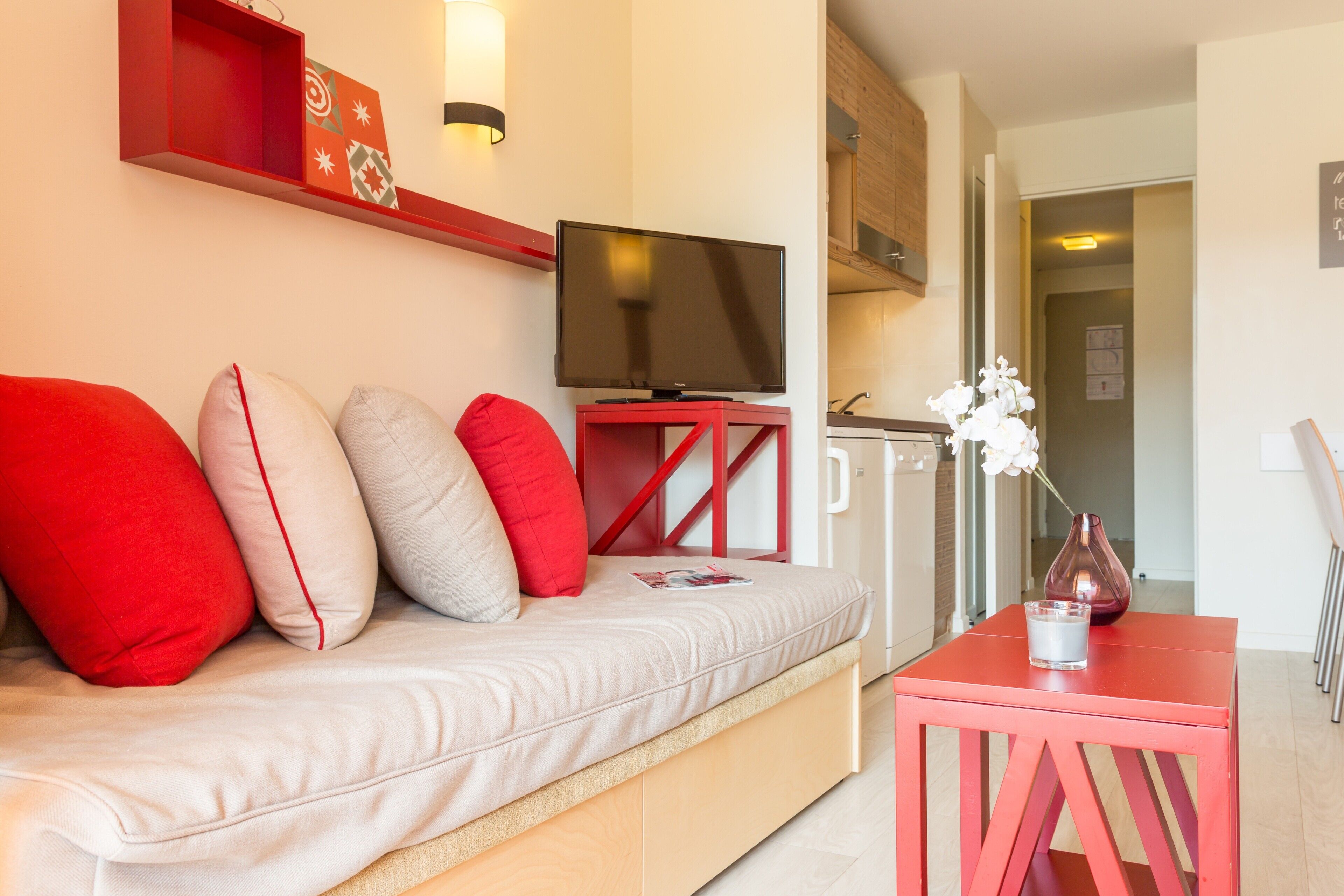 A modern living room with a sofa, red cushions, a flat-screen TV, a small table with a vase, and a red shelf with decorative items.