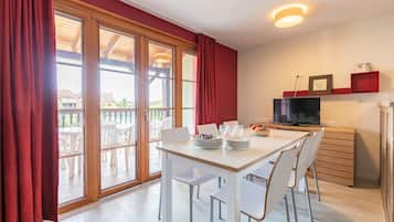 Een eethoek met een witte tafel en stoelen, een televisie op een dressoir, en een rode plank met een fotolijst.