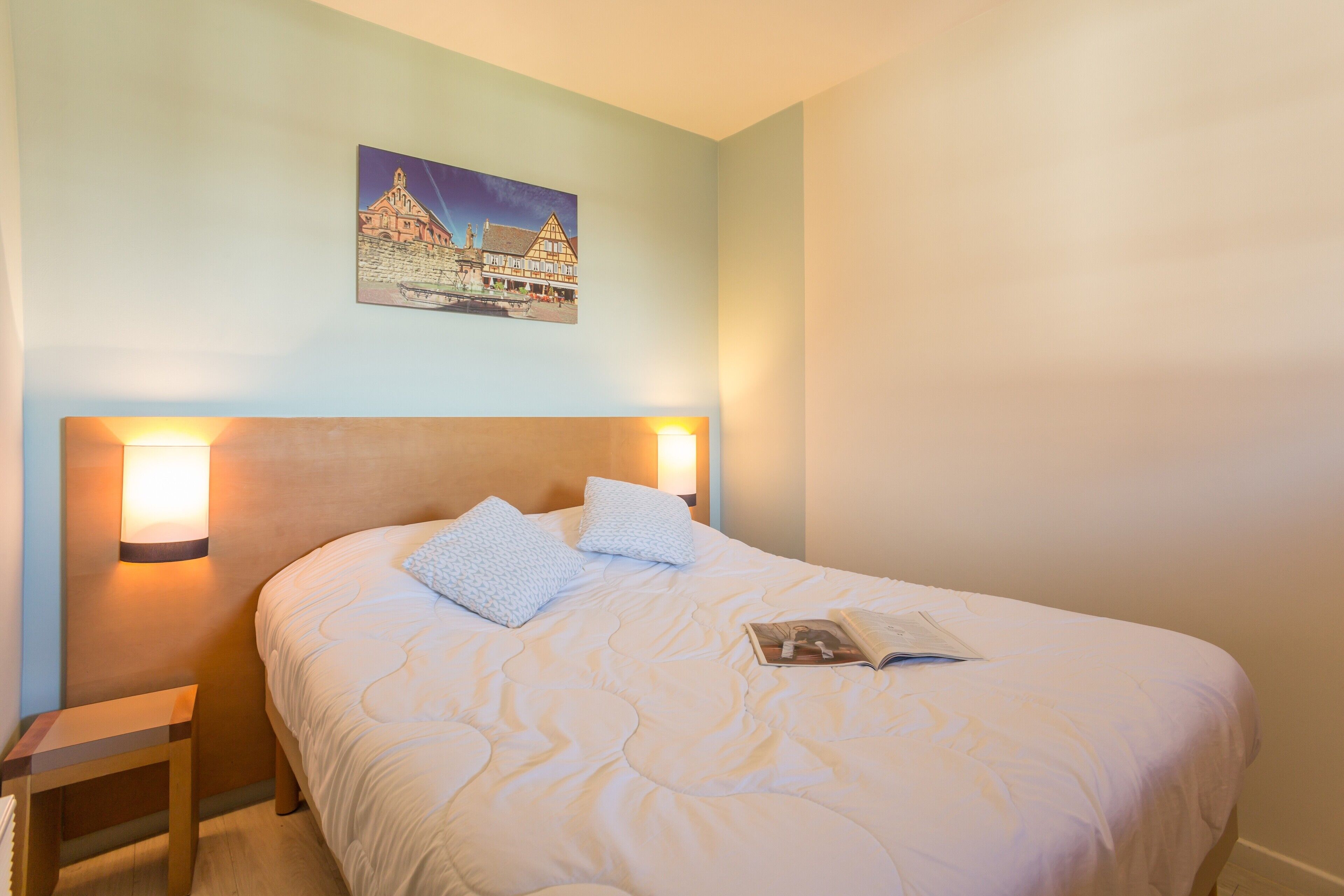 A neatly made bed with a quilted cover, two pillows, and a magazine on it. There is a small bedside table with a lamp beside the bed. A painting of a building is hung on the wall above the bed.