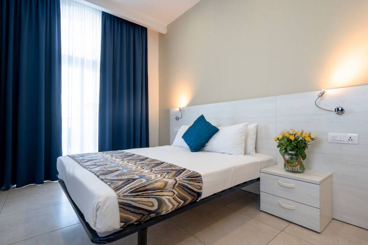 Een hotelkamer met een bed, een nachtkastje, een vaas met bloemen en een wandlamp.