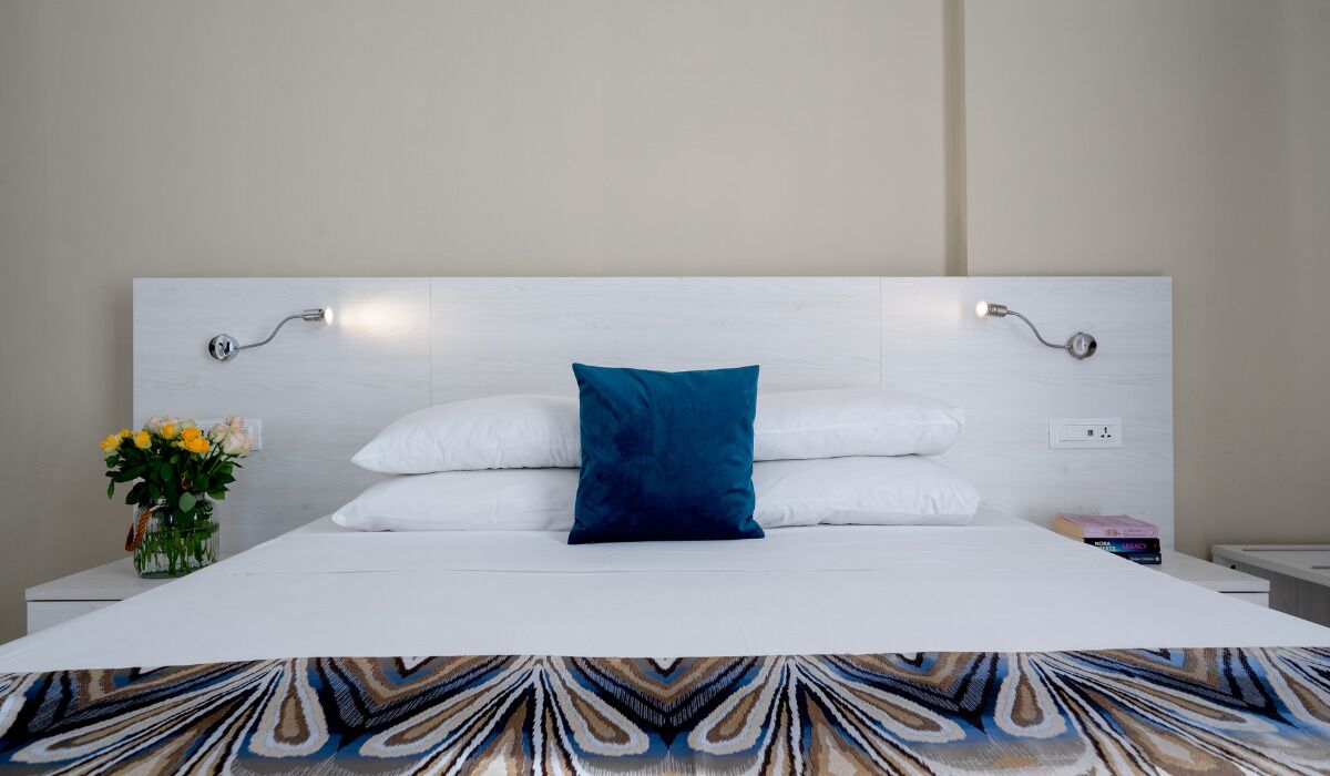 Una cama bien hecha con sábanas blancas y una almohada azul, una mesita de noche con un florero y una lámpara fijada a la pared.