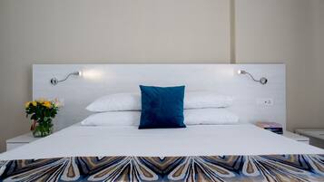 Una cama bien hecha con sábanas blancas y una almohada azul, una mesita de noche con un florero y una lámpara fijada a la pared.
