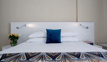 Una cama bien hecha con sábanas blancas y una almohada azul, una mesita de noche con un florero y una lámpara fijada a la pared.
