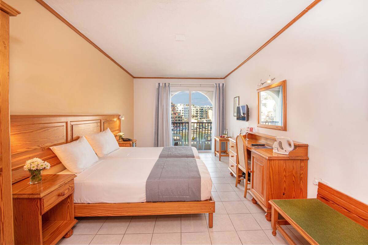 Een hotelkamer met een groot bed, houten meubels, een klein bureau en uitzicht op gebouwen door een raam.