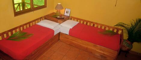 A room with two wooden beds, each with a red bedspread and a white pillow. There is a small wooden nightstand with a lamp and a framed picture. A potted plant is on the floor to the right.