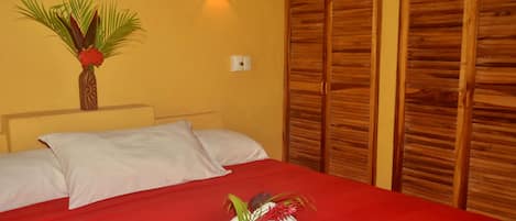 A hotel room with a red bedspread, white pillows, a wooden wardrobe, and a wall-mounted light fixture.