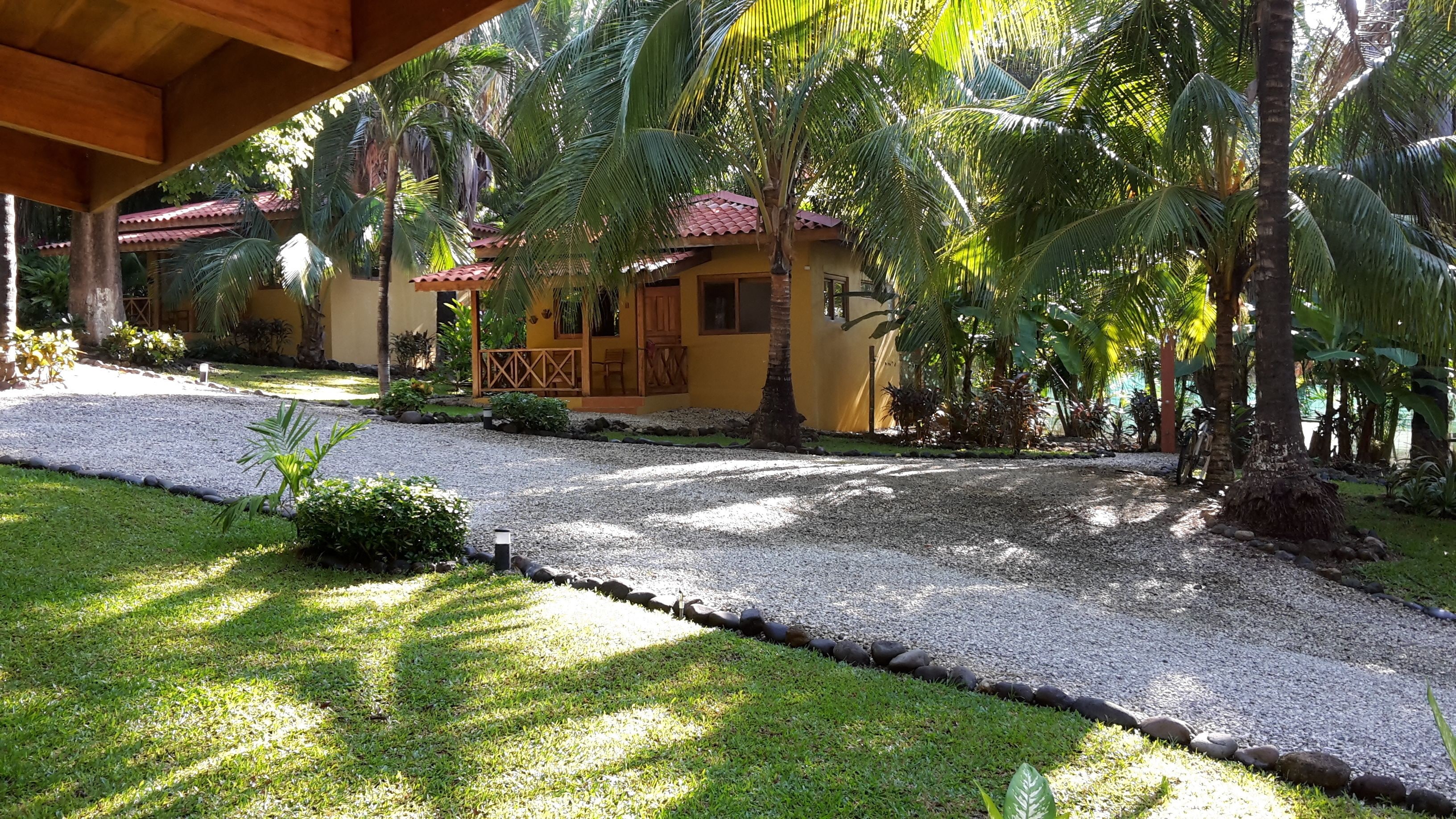 A tropical resort with a gravel pathway, palm trees, and a yellow building with a red roof.