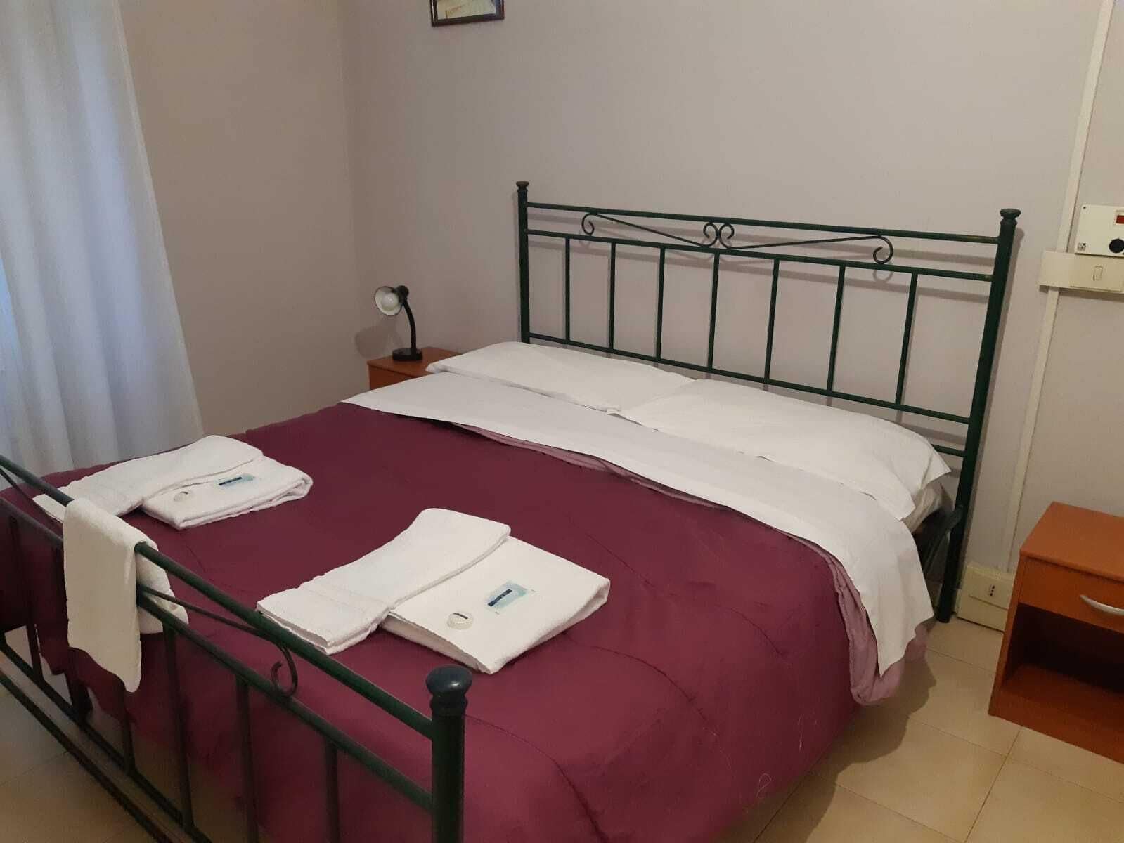 Un letto singolo con struttura in metallo, un cuscino bianco e una coperta rossa. È presente un comodino con una lampada e una presa di corrente.