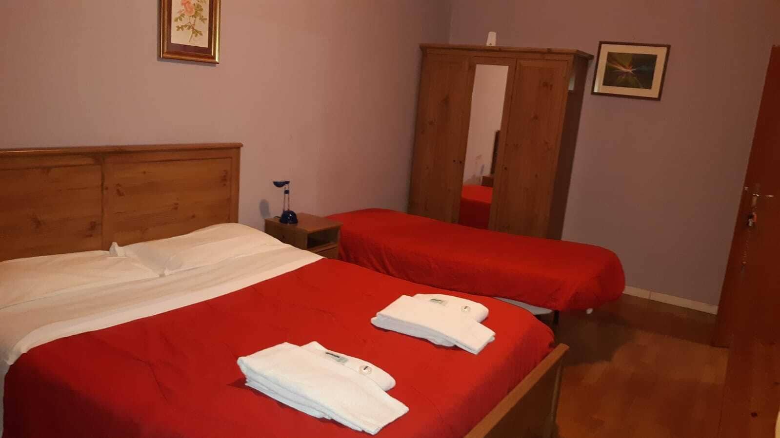 A room with two beds, each with a red bedspread and white towels. There is a wooden wardrobe and a small table with a lamp.