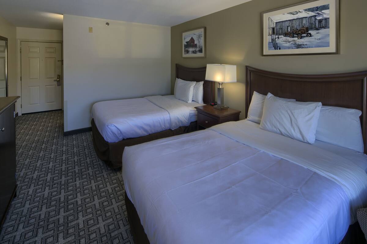 A hotel room with two beds, a nightstand with a lamp, a dresser, and a door leading to another room.