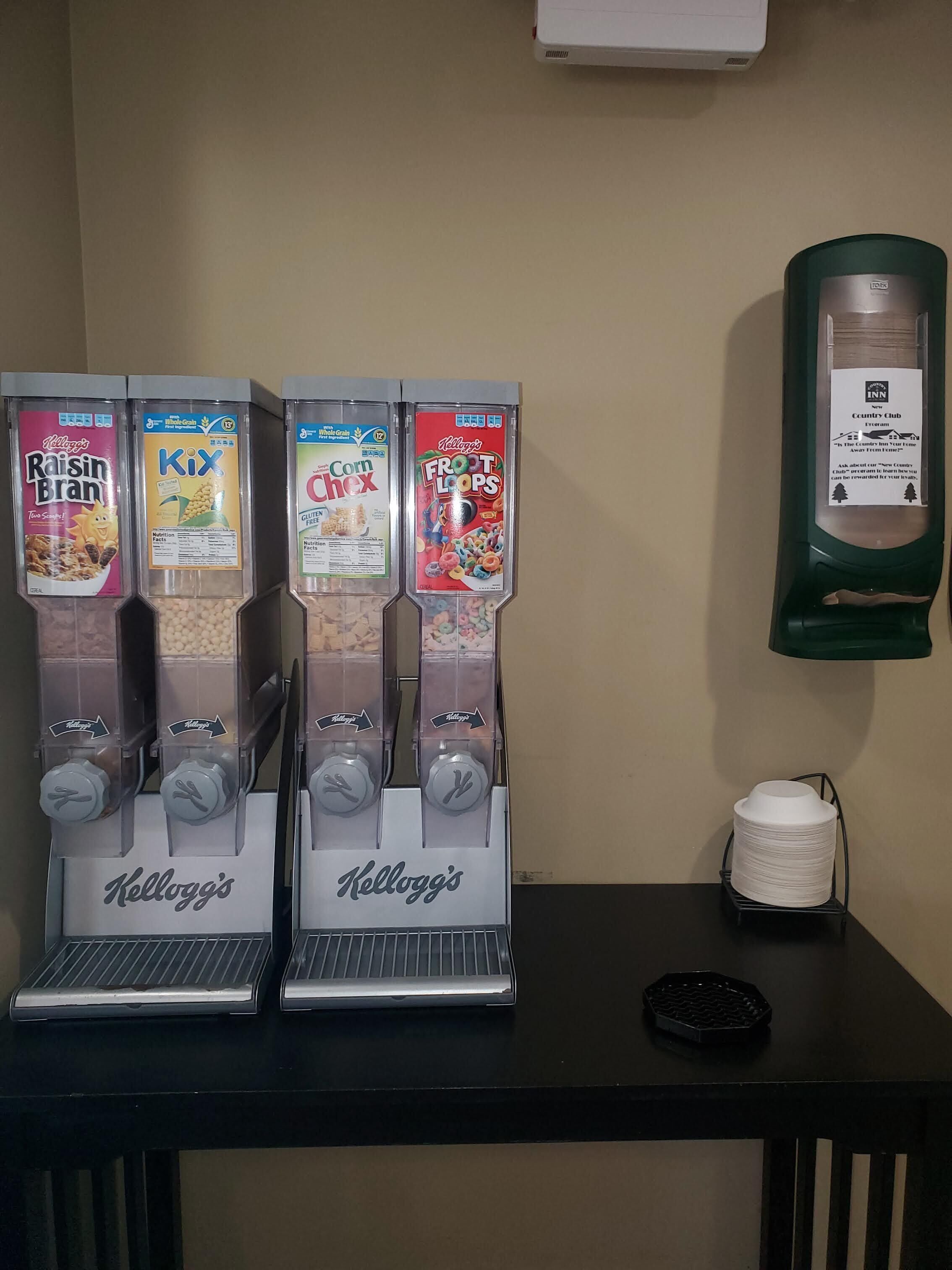 A row of Kellogg's cereal dispensers on a table with a green wall and a sign in the background.