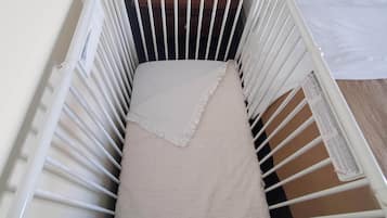 En hvid vugge med et beige tæppe og pude, placeret op ad en væg med en seng og puder i baggrunden.