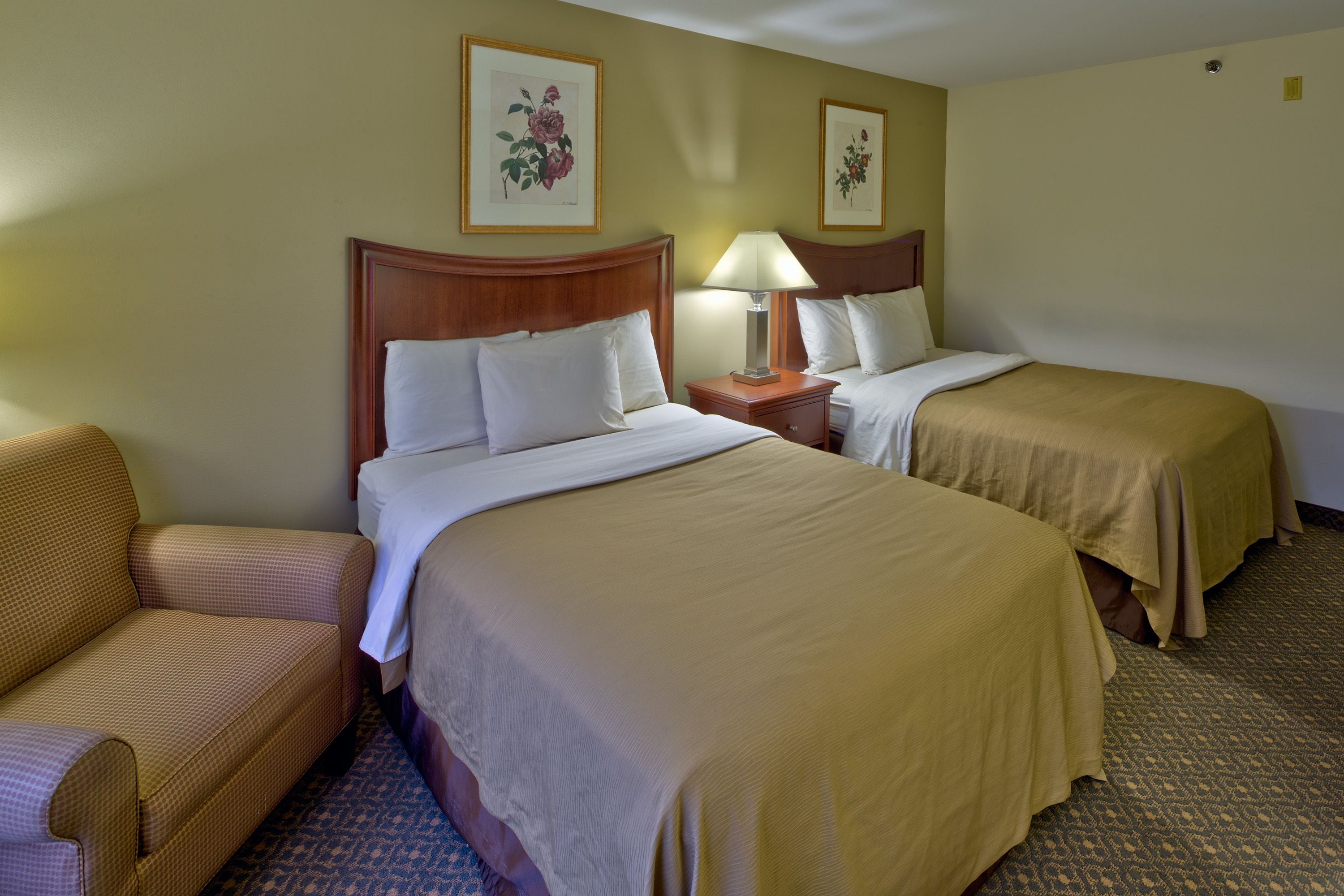 A hotel room with two beds, a nightstand with a lamp, a chair, and two framed floral pictures on the wall.