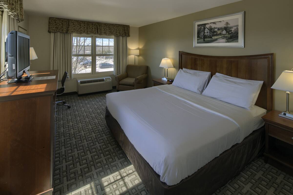 A hotel room with a large bed, a desk with a computer, a chair, a window with curtains, and a painting on the wall.