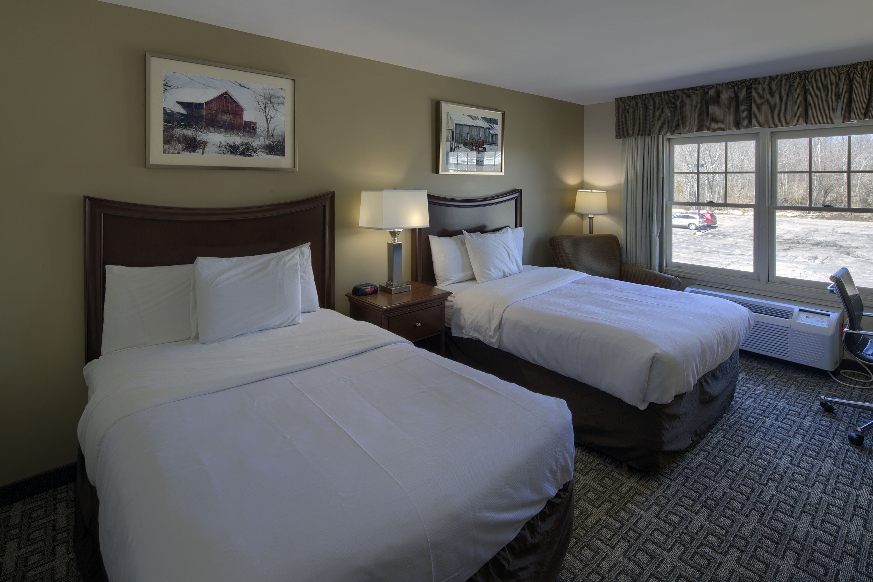 A hotel room with two beds, a window with a view of a parking lot, and a heater on the floor.
