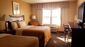 A hotel room with two beds, a desk with a computer, a TV, a window with curtains, and a framed picture on the wall.