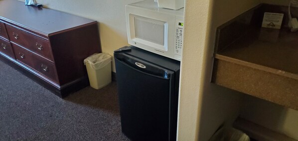 A kitchenette with a microwave, a black refrigerator, a trash bin, and a wooden dresser.