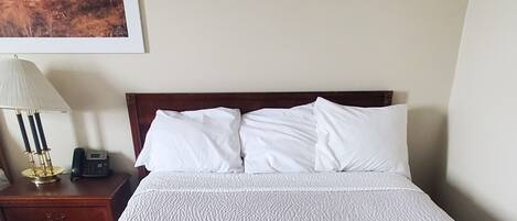 A neatly made bed with white bedding, a bedside table with a lamp and phone, and a painting on the wall.