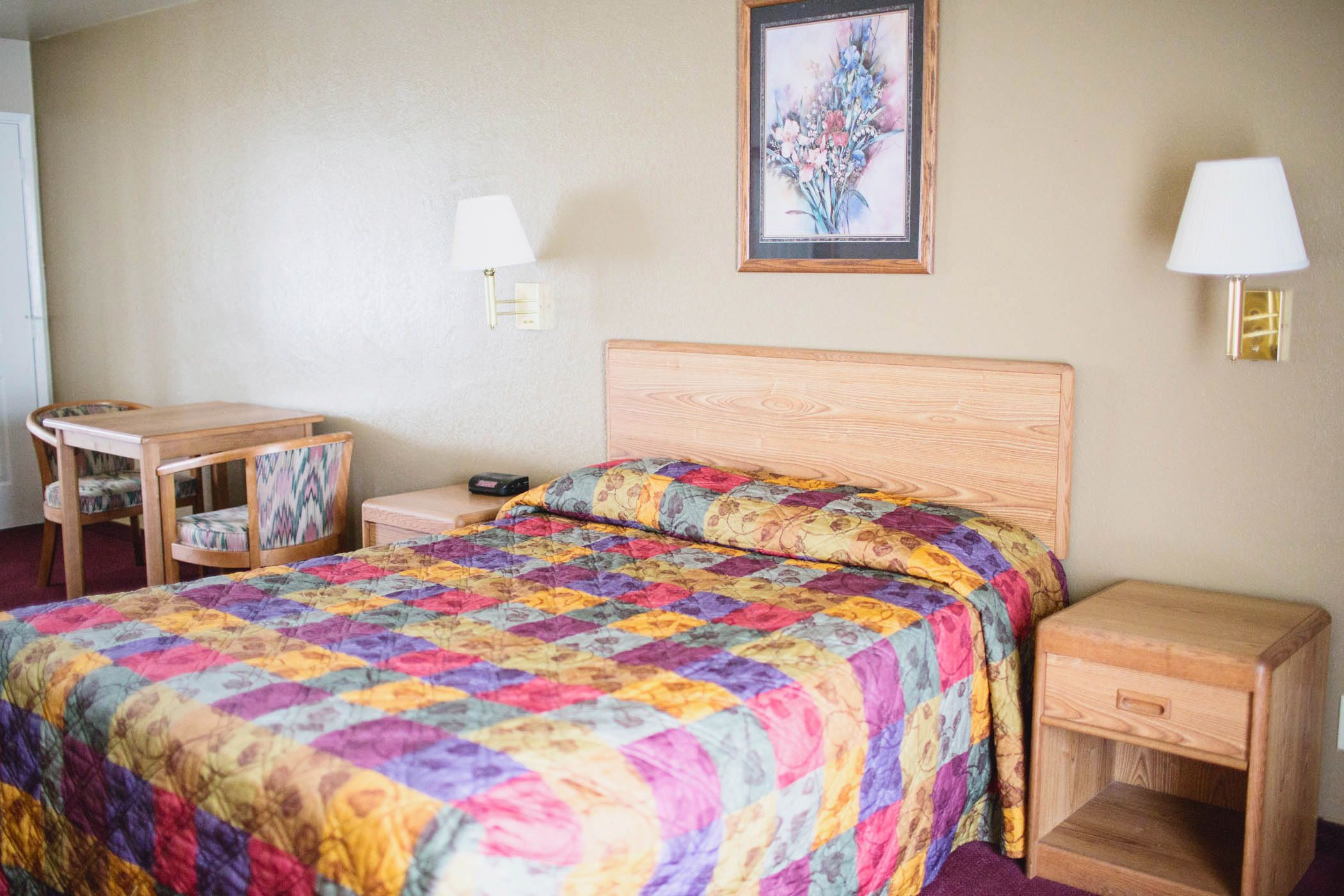A bedroom with a wooden bed, a bedside table, a chair, a desk, and a wall-mounted lamp.
