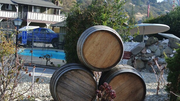 A pool area with wooden barrels, rocks, and a building with a pool in the background.