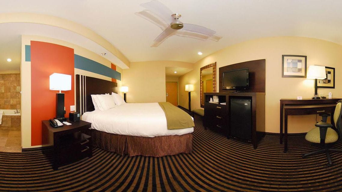 A hotel room with a bed, a desk, a chair, a television, a mirror, and a ceiling fan.