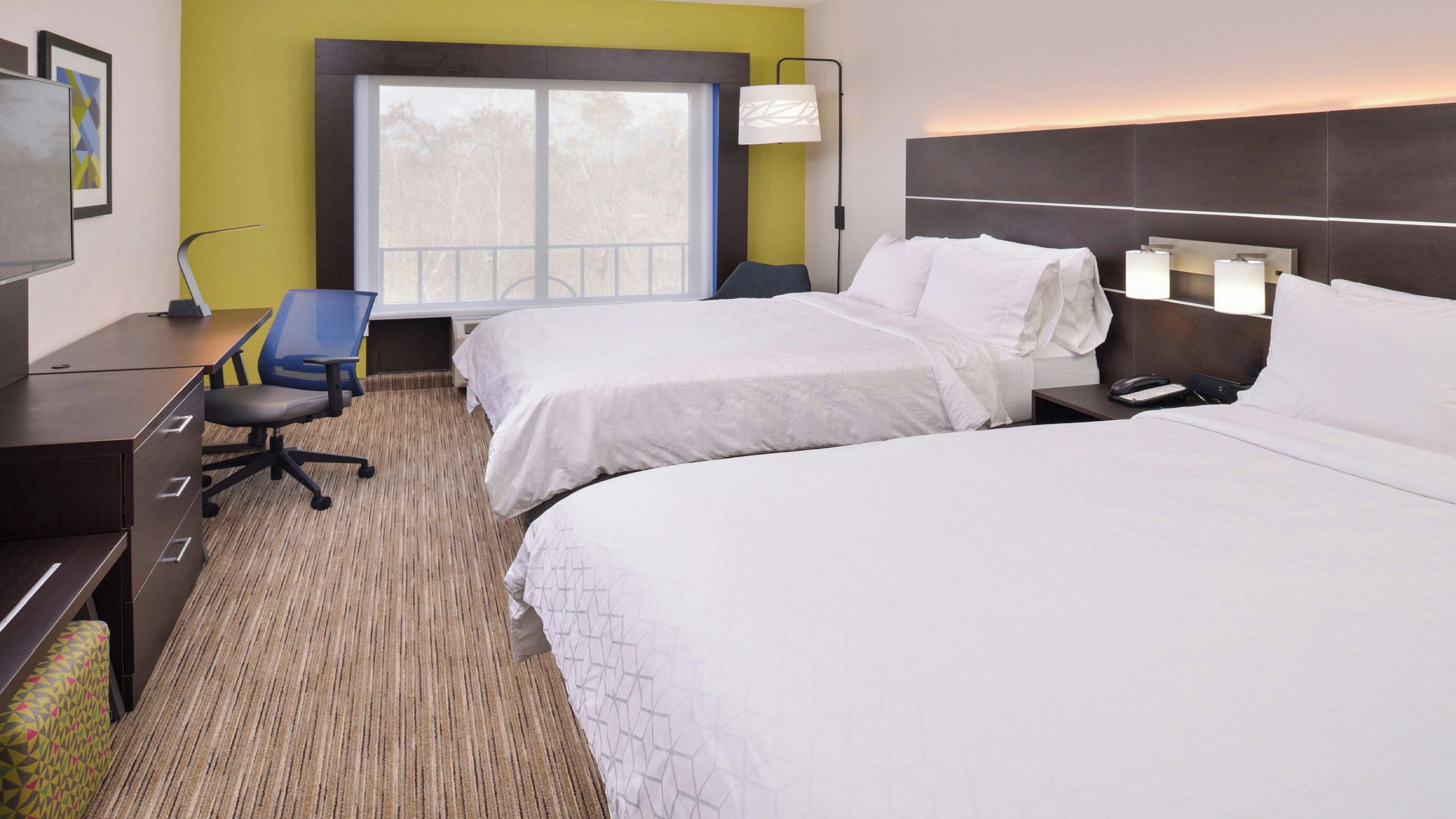 A hotel room with two beds, a desk, a chair, a lamp, and a window.