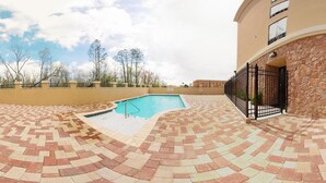 A swimming pool with a tiled deck, surrounded by a low stone wall and a building with a black metal fence. - Holiday Inn Express & Suites Laplace by IHG (LaPlace)