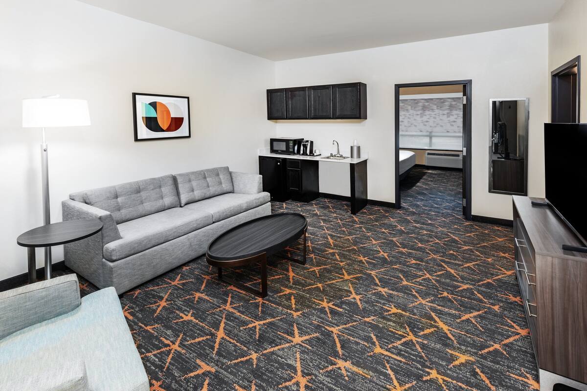A hotel room with a grey sofa, a small round table, a kitchenette, a TV, and a patterned carpet.