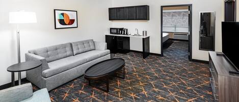 A hotel room with a grey sofa, a small round table, a kitchenette, a TV, and a patterned carpet.
