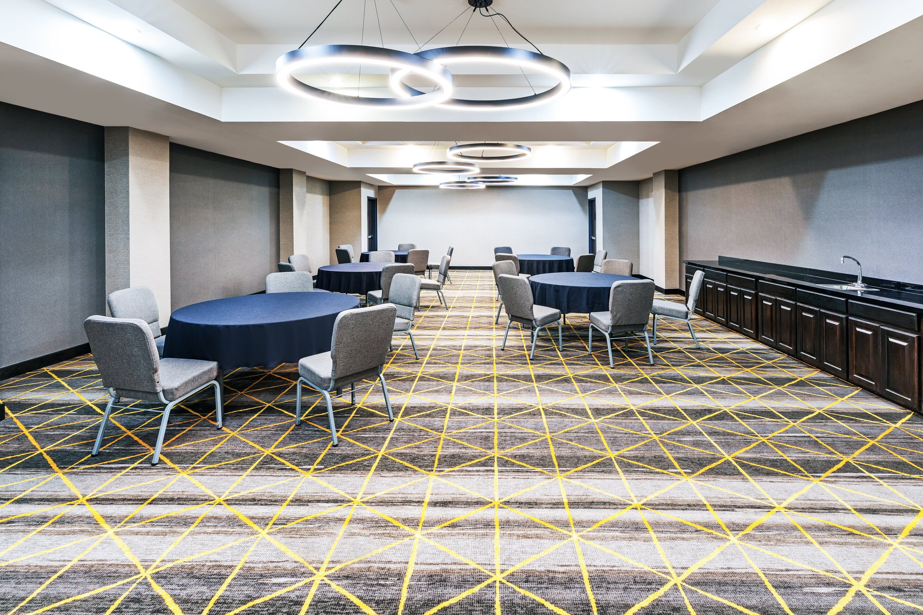 Un salón de conferencias con mesas y sillas redondas, una alfombra estampada y una zona de cocina con fregadero.
