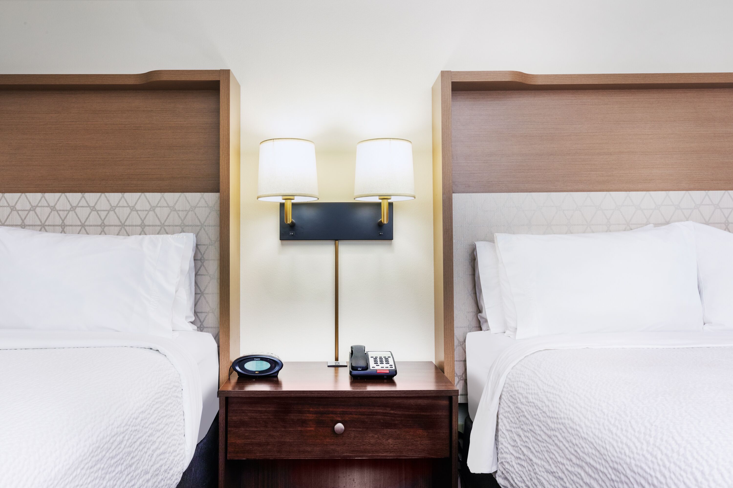 Een hotelkamer met twee bedden, een nachtkastje met telefoon en lamp, en een wandlamp.