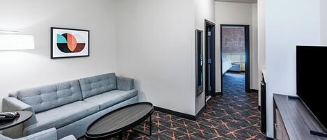 Habitación de hotel con sofá gris, una mesita redonda pequeña, televisor de pantalla plana y alfombra estampada.