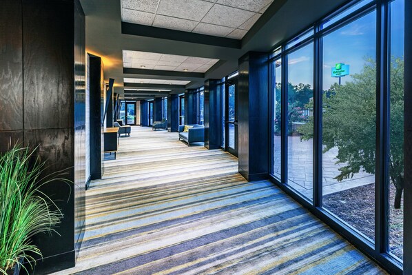 A hotel corridor with striped carpet, large windows, and a seating area with a blue couch and a chair. - Holiday Inn Stillwater - University West by IHG (Stillwater)