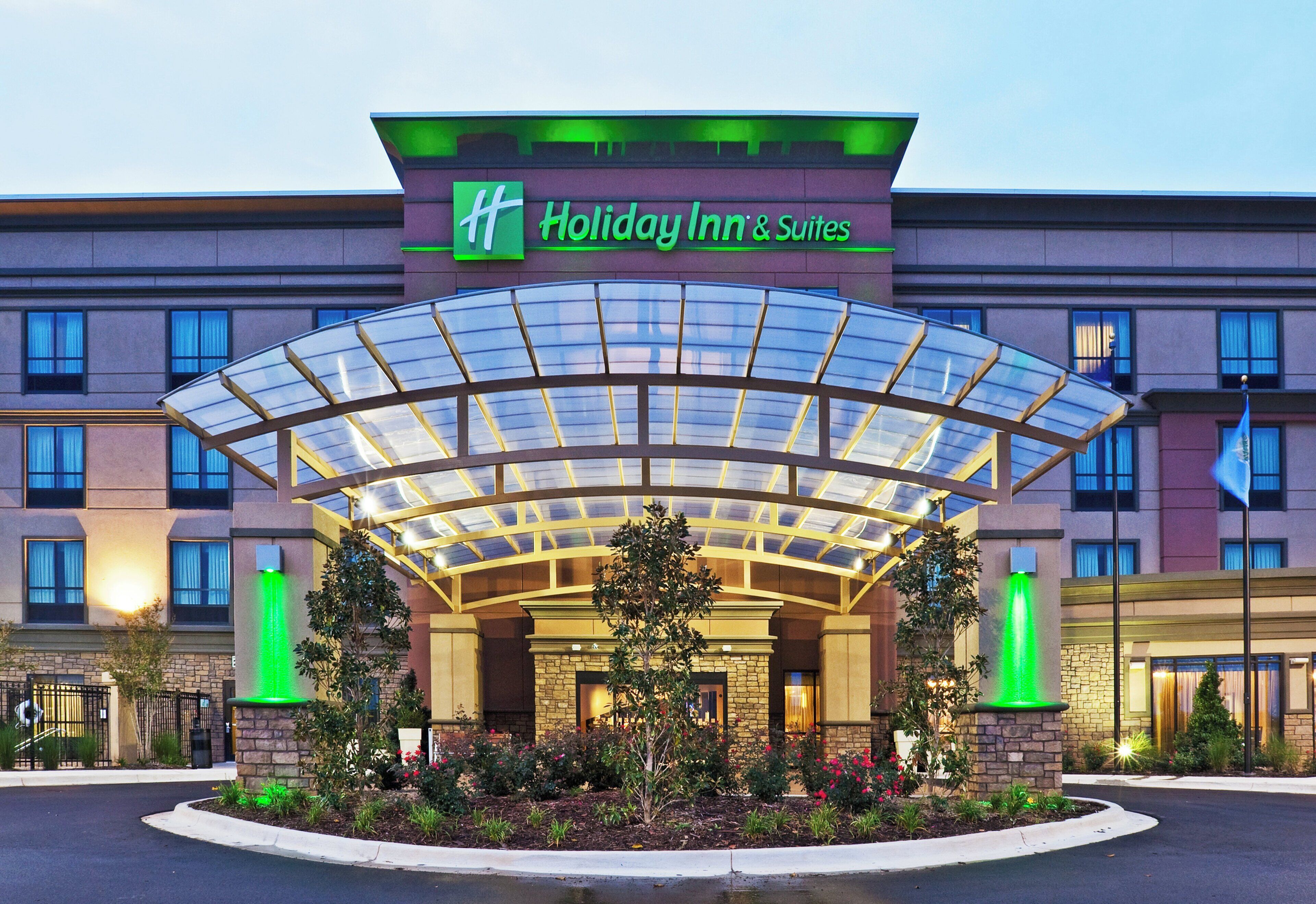 Un hôtel Holiday Inn & Suites doté d’un auvent d’entrée en verre, d’une façade en pierre et d’un espace d’entrée aménagé.