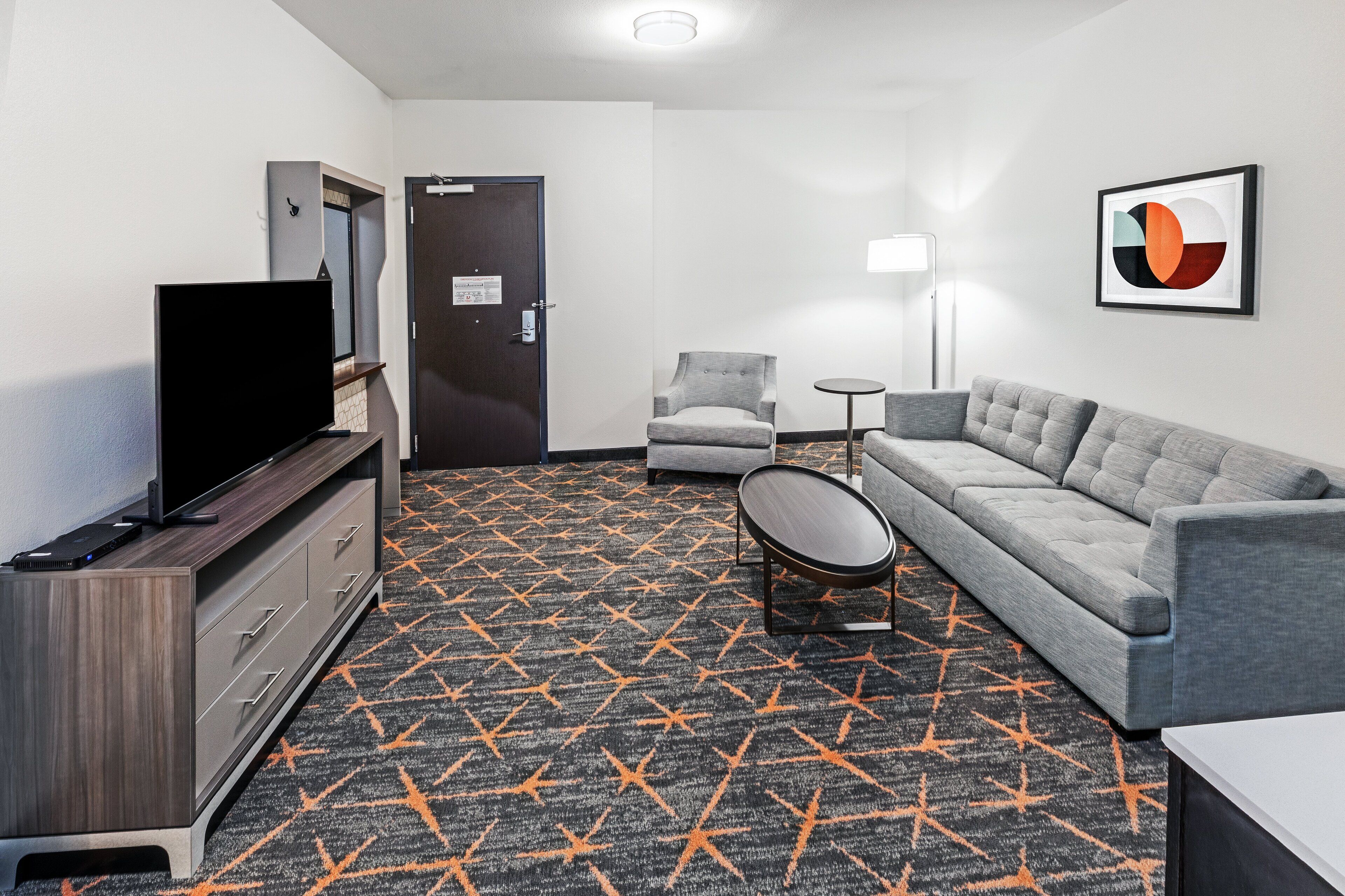 A hotel room with a flat-screen TV, a sofa, a chair, a small table, and a patterned carpet.