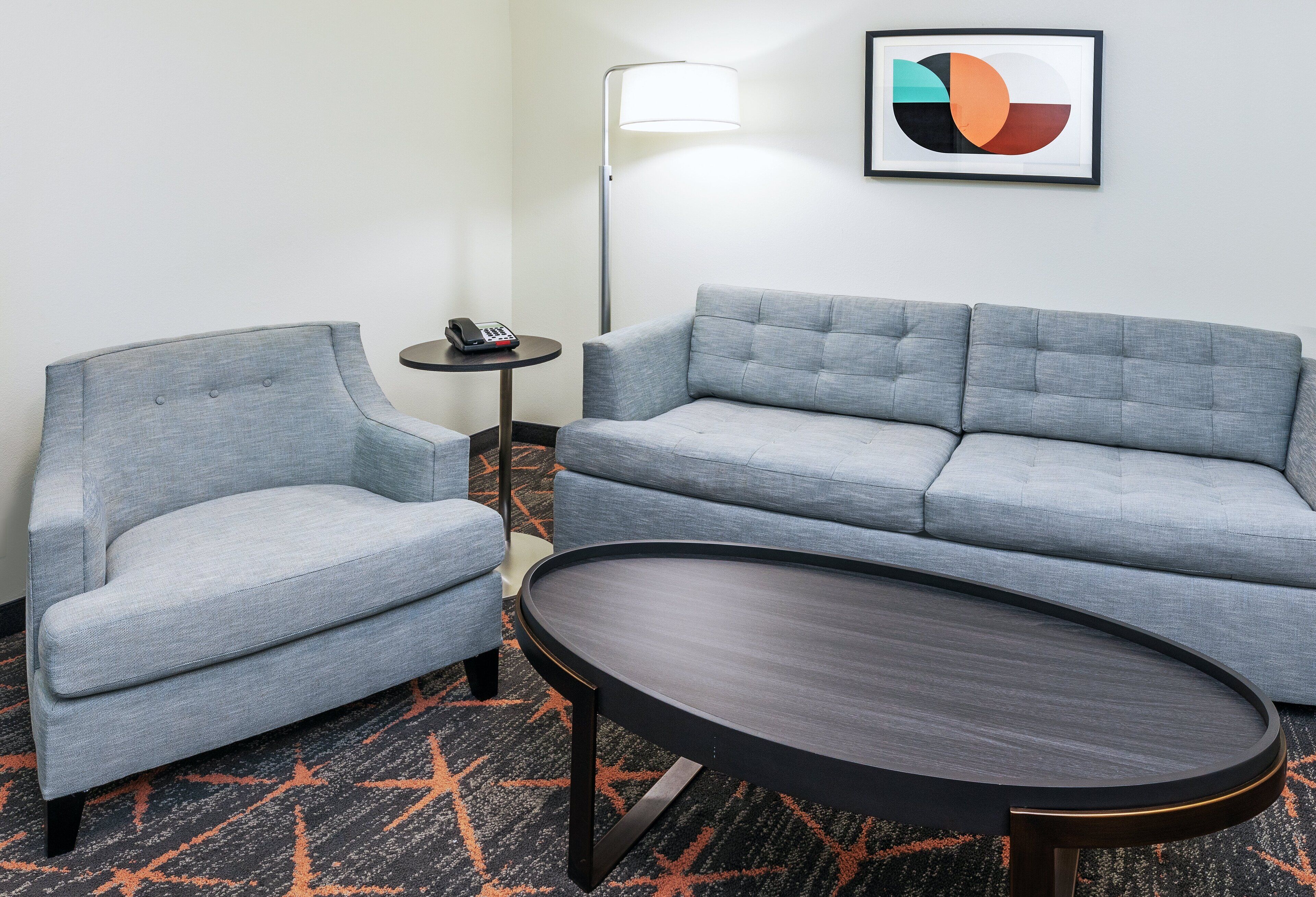 A modern hotel room with a grey sofa, a matching armchair, a round coffee table, a small side table with a phone, and a floor lamp.