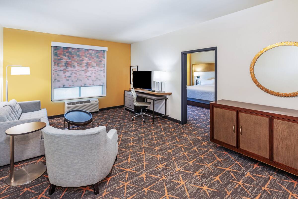 A hotel room with a bed, a desk, a chair, a small table, a TV, a lamp, and a mirror.