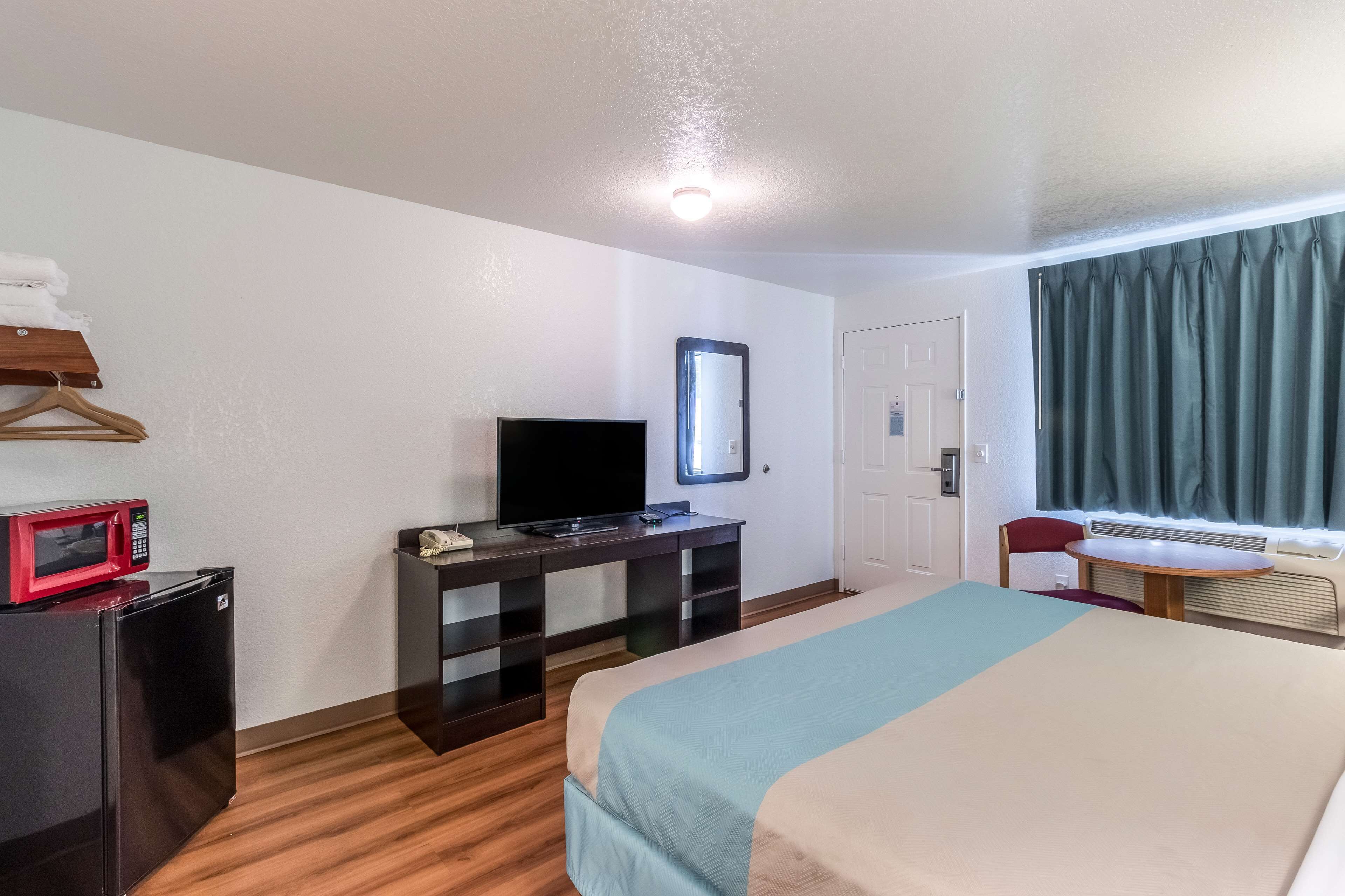 Habitación de hotel con cama, escritorio con televisor, microondas, refrigerador pequeño, espejo, ventana con cortinas y puerta.